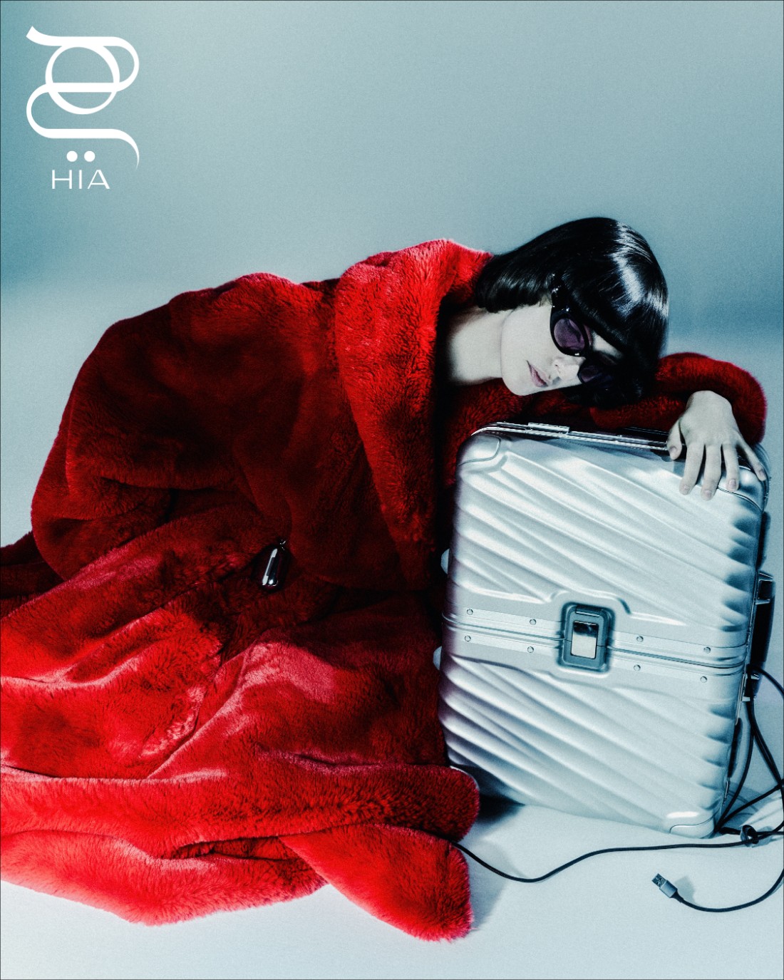 من "بالنسياغا" Balenciaga، معطف أحمر من الفرو الزائف، وحذاء من جلد العجل باللون الأسود اللامع. حقيبة سفر من "تومي" Tumi، ونظارات شمسية من "جينتل مونستر" Gentle Monster.