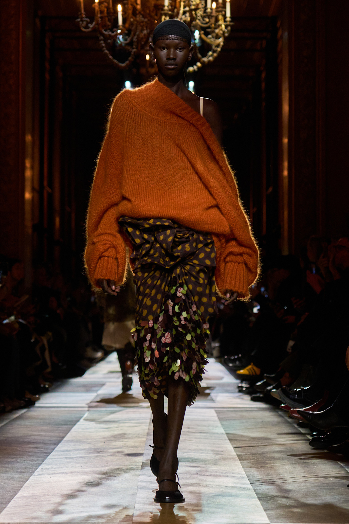 تنورة مرقطة مع تطريز براق من Dries Van Noten نُسّقت مع كنزة صوفية كبيرة باللون البرتقالي، في مزيج يعكس روحًا فنية غير تقليدية وأناقة عفوية