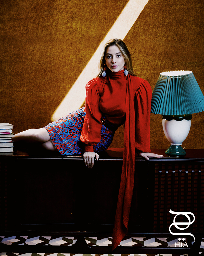 قميص من الحرير والجاكار باللون الأحمر الكركديه ومزين بنقشة الورود تنورة ميدي من الصوف ومطرزة بدانتيل زهري باللونين الأزرق والأحمر حزام من جلد الأفعى باللون الأسود مع مشبك مربّع بحرف G أقراط بتأثير حجر الراتينج باللون المرجاني جميعها من دار "غوتشي" GUCCI