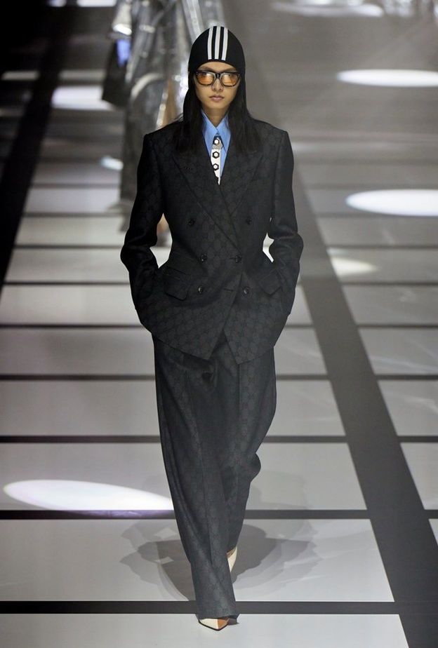 بذلة رمادية، قميص أزرق فاتح، قبّعة سوداء ذات خطوط بيضاء، وربطة عنق مزخرفة باللونين الأبيض والرمادي من غوتشي