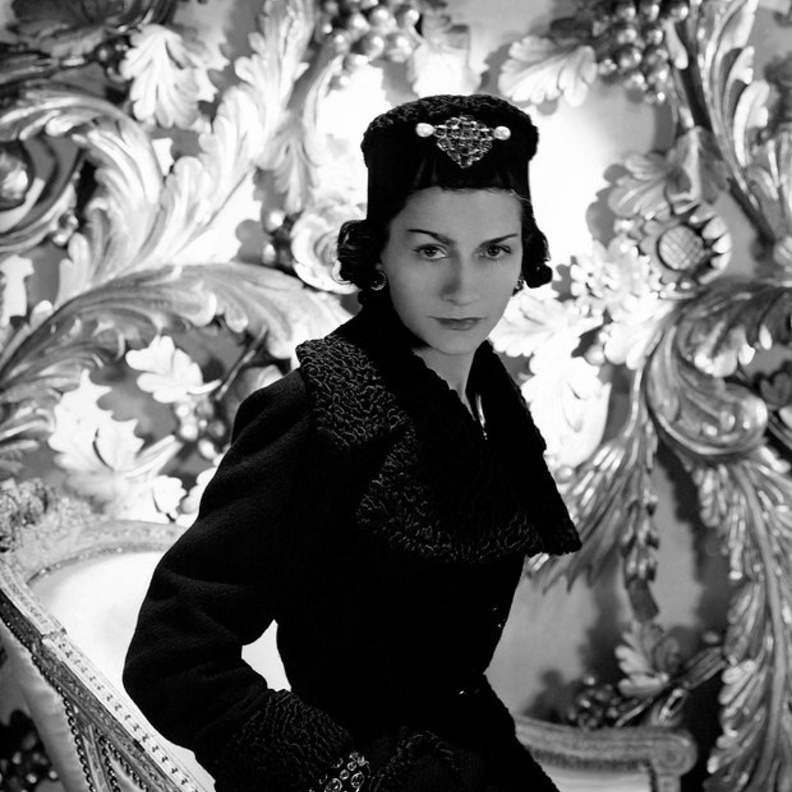 صورة لـ كوكو شانيل وهي ترتدي ملابس من تصميمها عبارة عن معطف باللون الأسود وقبعة مزينة ببروش مميز