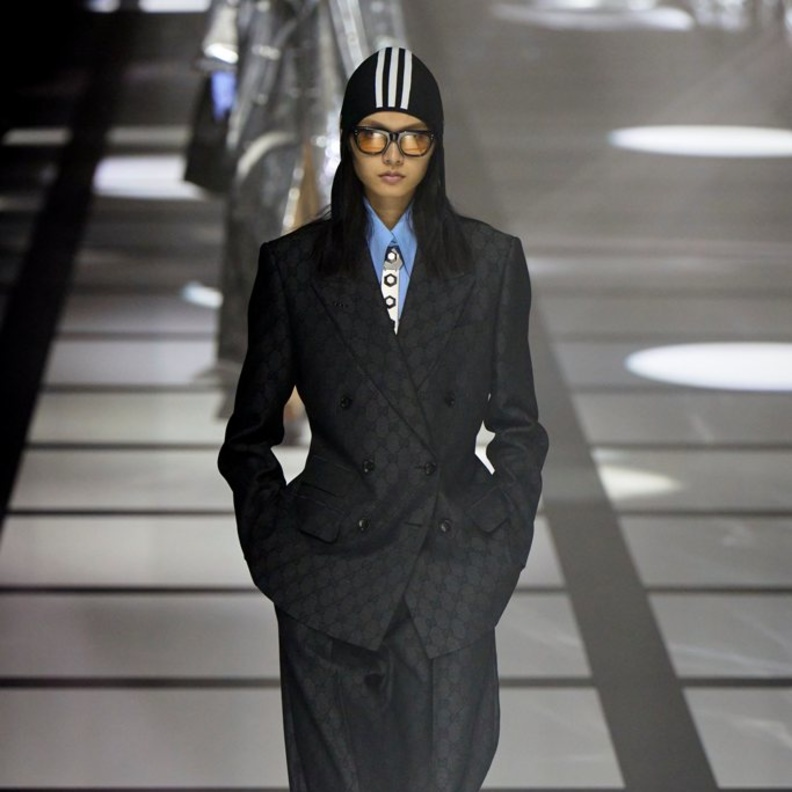 بذلة رمادية، قميص أزرق فاتح، قبّعة سوداء ذات خطوط بيضاء، وربطة عنق مزخرفة باللونين الأبيض والرمادي من غوتشي